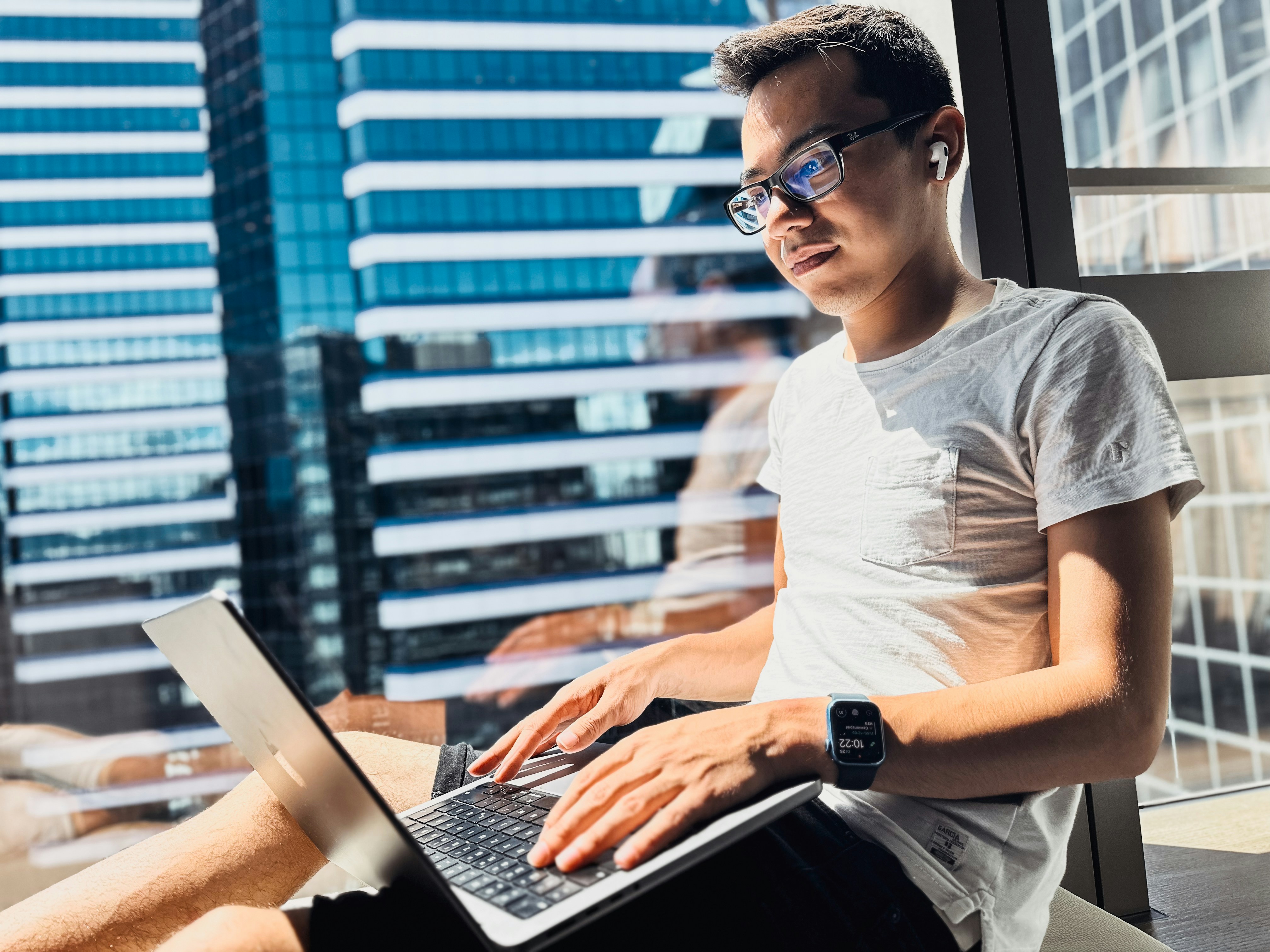 A young digital native is sitting on a window sill, wearing glasses and earbuds. He has his legs crossed and is using a laptop computer that rests on his lap. The sun shines through the window behind him, illuminating his face with its warm light. In background the skyline of Quarry Bay, Hong Kong.